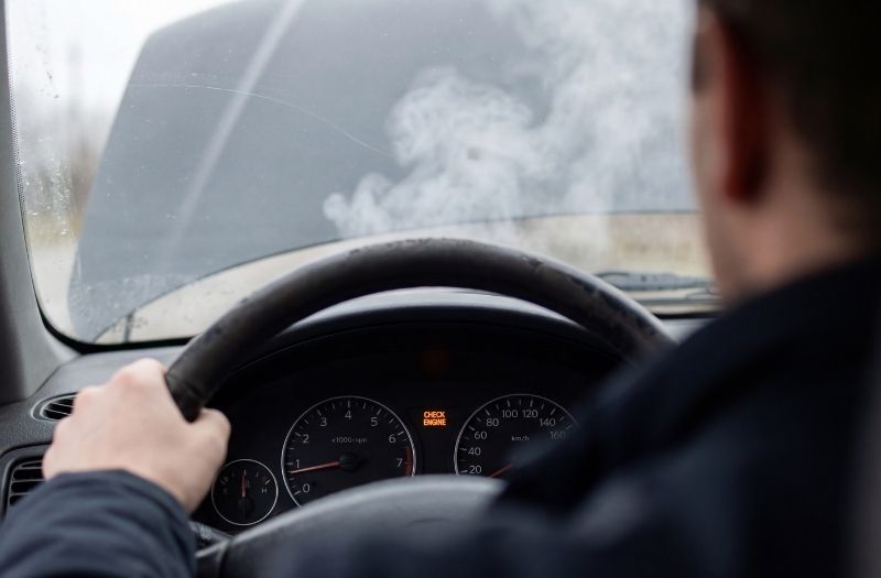 Driver view of broken down car with smoke rising from open hood and check engine light on dashboard due to engine overheating.